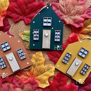 Pu&ograve; includere: Tre ornamenti a forma di casa in verde scuro, marrone e giallo senape. Ognuno presenta una porta chiara con un piccolo cuore, tre finestre con vetri bianchi e puntini bianchi. Ogni ornamento &egrave; contrassegnato con le lettere HBS.