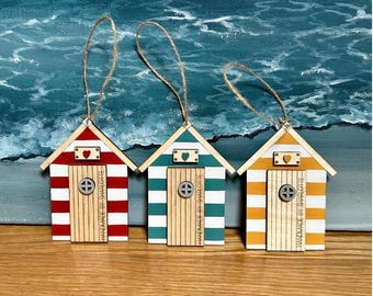 Pode incluir: Três ornamentos de madeira em forma de cabana de praia com cordões de juta para pendurar. Cada cabana apresenta riscas horizontais vermelhas, azul-petróleo ou amarelas, uma porta central de madeira com janela circular e um coração.