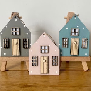 Pu&ograve; includere: Tre ornamenti in legno a forma di casa nei colori grigio, rosa chiaro e blu, ciascuno con pois bianchi, una porta in legno chiaro con pomello a forma di cuore e tre finestre. Ogni casa &egrave; esposta su un piccolo cavalletto in legno.