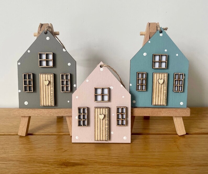 Pu&ograve; includere: Tre ornamenti in legno a forma di casa nei colori grigio, rosa chiaro e blu, ciascuno con pois bianchi, una porta in legno chiaro con pomello a forma di cuore e tre finestre. Ogni casa &egrave; esposta su un piccolo cavalletto in legno.
