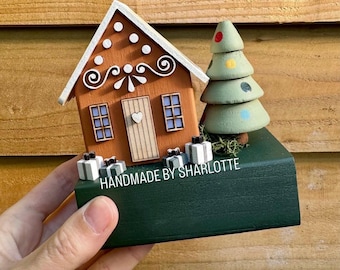 Può includere: Un ornamento in legno a forma di casa di pan di zenzero con facciata marrone, tetto bianco, porta centrale con cuore e base verde. Include un albero verde stilizzato con pois colorati e piccoli pacchetti regalo. Testo: HANDMADE BY SHARLOTTE.