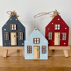 Pu&ograve; includere: Tre ornamenti in legno a forma di casa in grigio scuro, azzurro e rosso, ciascuno con pois bianchi, una porta in legno chiaro con pomello a forma di cuore e finestre. Le porte recano la scritta HANDMADE BY SHARLOTTE.