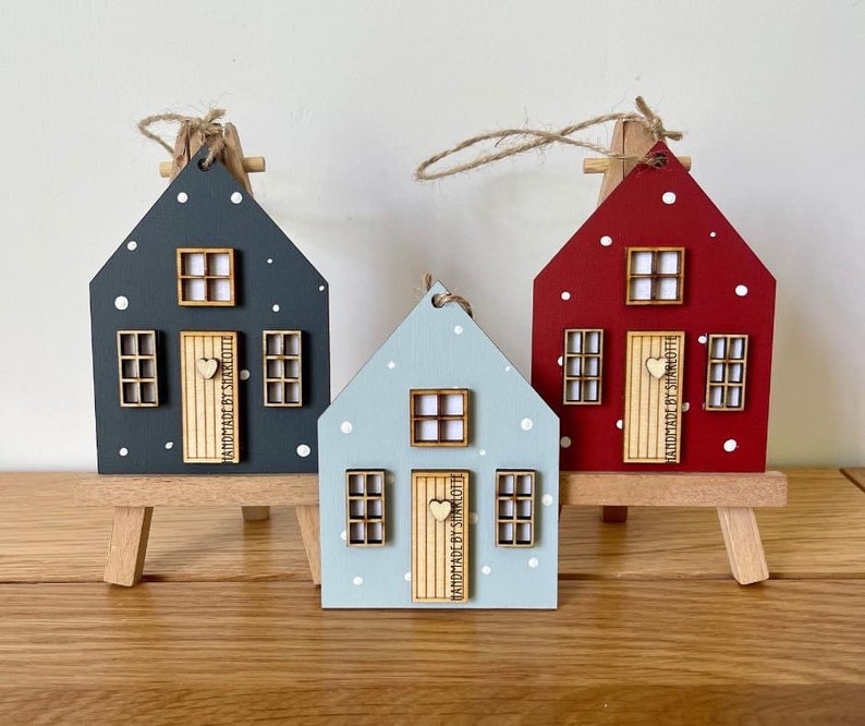 Pu&ograve; includere: Tre ornamenti in legno a forma di casa in grigio scuro, azzurro e rosso, ciascuno con pois bianchi, una porta in legno chiaro con pomello a forma di cuore e finestre. Le porte recano la scritta HANDMADE BY SHARLOTTE.
