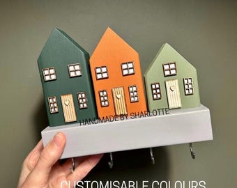 Può includere: Un portachiavi da parete con tre ornamenti in legno a forma di casa nei colori verde scuro, arancione e verde chiaro. Ogni casa presenta un piccolo cuore sulla porta e finestre quadrate. La base è una mensola rettangolare grigio chiaro con quattro ganci.