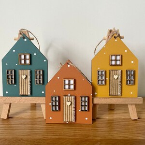 Pu&ograve; includere: Tre ornamenti in legno a forma di casa nei colori verde acqua, terracotta e giallo senape, con pois bianchi, una piccola porta in legno con un cuore e finestre a griglia. Ogni porta riporta la scritta verticale Handmade by Sharlotte.