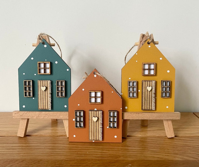 Pu&ograve; includere: Tre ornamenti in legno a forma di casa nei colori verde acqua, terracotta e giallo senape, con pois bianchi, una piccola porta in legno con un cuore e finestre a griglia. Ogni porta riporta la scritta verticale Handmade by Sharlotte.