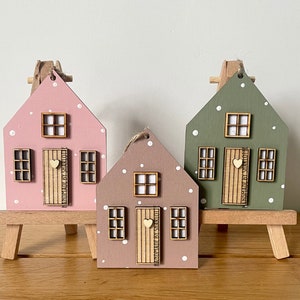 Pu&ograve; includere: Tre ornamenti in legno a forma di casa nei colori rosa, malva e verde salvia, ciascuno con pois bianchi, una porta centrale con un piccolo cuore e tre finestre. Ogni porta riporta la scritta HANDMADE BY SHARLOTTE.
