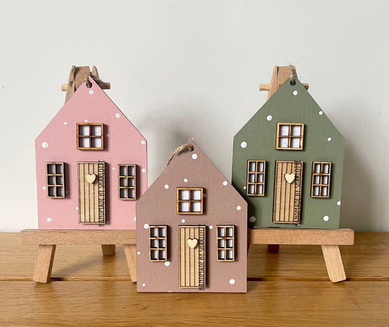 Pu&ograve; includere: Tre ornamenti in legno a forma di casa nei colori rosa, malva e verde salvia, ciascuno con pois bianchi, una porta centrale con un piccolo cuore e tre finestre. Ogni porta riporta la scritta HANDMADE BY SHARLOTTE.
