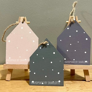 Pu&ograve; includere: Tre ornamenti in legno a forma di casa nei colori rosa chiaro, grigio scuro e viola, decorati con pois bianchi e un laccio in iuta. Il testo "HANDMADE BY SHARLOTTE" &egrave; stampato sulla base di ogni pezzo, esposto su piccoli cavalletti in legno.