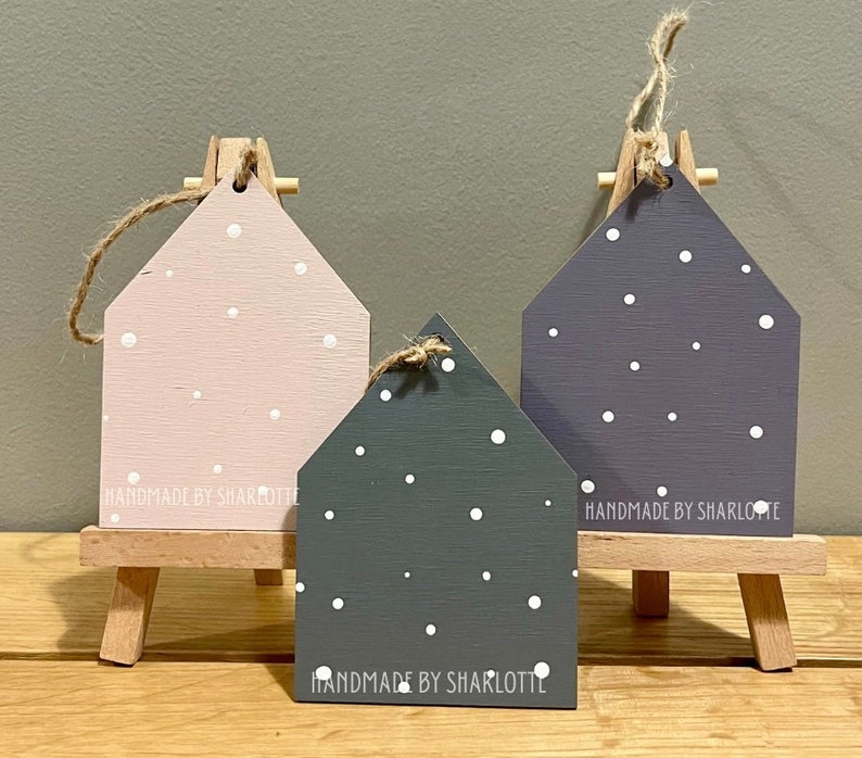 Pu&ograve; includere: Tre ornamenti in legno a forma di casa nei colori rosa chiaro, grigio scuro e viola, decorati con pois bianchi e un laccio in iuta. Il testo "HANDMADE BY SHARLOTTE" &egrave; stampato sulla base di ogni pezzo, esposto su piccoli cavalletti in legno.