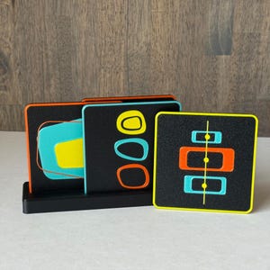 May include: Set of square coasters with a black background and colorful geometric designs. The coasters feature orange, yellow, and teal shapes. They are displayed in a black holder, showcasing a retro, mid-century modern style.