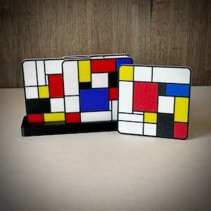 May include: A set of two coasters with a Mondrian-inspired design. The coasters feature a black border with a white background and colorful geometric shapes in red, yellow, and blue. One coaster is in a black stand.