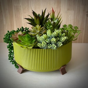 May include: A round, lime green planter with vertical ribbed detailing, holding a variety of artificial succulents. The planter has three brown, angled legs. The succulents are various shades of green, with some red accents.