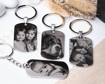Puede incluir: Cuatro llaveros de metal plateado con forma de placa de identificación, cada uno con una fotografía distinta grabada con láser en blanco y negro. Las imágenes incluyen retratos de dos niños, un perro, una familia y un bebé.
