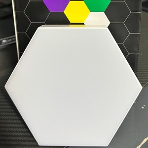 Puede incluir: Una luz LED hexagonal blanca con una caja negra detrás. La caja tiene un patrón de panal y el texto "Hexagonal LED light". La caja también muestra un patrón hexagonal de luces de colores en rojo, naranja, amarillo, verde, azul y rosa.