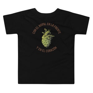 Może przedstawiać: Czarny t-shirt dla malucha z grafiką serca z kaktusa. Tekst wokół serca brzmi "CON EL NOPAL EN LA FRENTE Y EN EL CORAZÓN". Serce z kaktusa jest zielone i brązowe.
