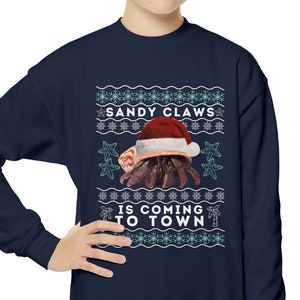 May include: A navy blue sweatshirt with a Christmas-themed design featuring a hermit crab wearing a Santa hat. The text on the sweatshirt reads "Sandy Claws is coming to town".