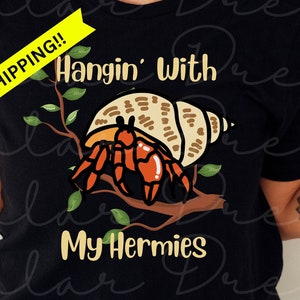 May include: Black t-shirt with a graphic of a hermit crab in a shell on a branch with green leaves. The text "Hangin' With My Hermies" is printed below the graphic. The text "FREE SHIPPING!" is printed in yellow on a white banner.