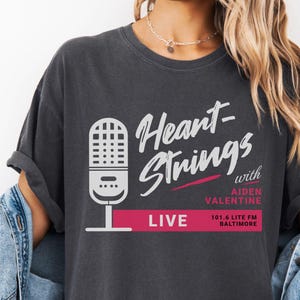 May include: Dark gray t-shirt featuring the text "Heart Strings with Aiden Valentine LIVE" and a microphone graphic. The shirt has a vintage look and is made of soft material. The shirt is part of the apparel category.