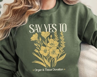 May include: A green sweatshirt with the text "SAY YES TO" and a floral design. Below the text is the phrase "*Organ & Tissue Donation*."