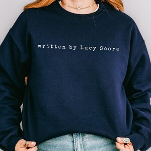 May include: A navy blue sweatshirt with the text "written by Lucy Score" in white on the front.