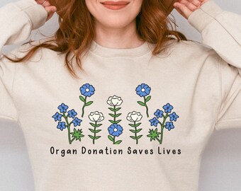 Puede incluir: Una sudadera beige con un gráfico de flores azules y blancas y el texto "Organ Donation Saves Lives".