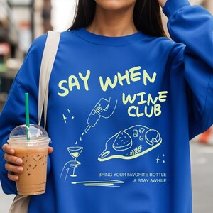 May include: A royal blue crewneck sweatshirt featuring a light yellow graphic print that reads "SAY WHEN WINE CLUB" and "BRING YOUR FAVORITE BOTTLE & STAY AWHILE," with illustrations of a wine bottle, a glass, and a charcuterie board with cheese.