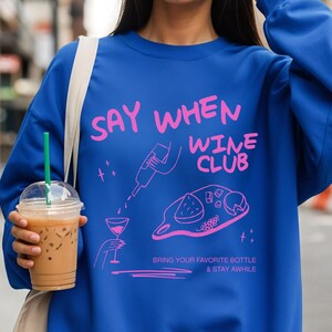 May include: A royal blue crewneck sweatshirt featuring a pink graphic print. The design includes the text "SAY WHEN WINE CLUB" and "BRING YOUR FAVORITE BOTTLE & STAY AWHILE" alongside line art of a wine bottle, a glass, and a charcuterie board.
