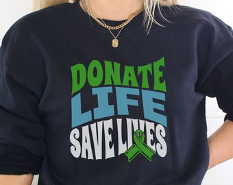 Puede incluir: Una sudadera negra con el texto "DONATE LIFE SAVE LIVES" en letras verdes, azules y blancas. Una cinta verde se presenta debajo del texto.