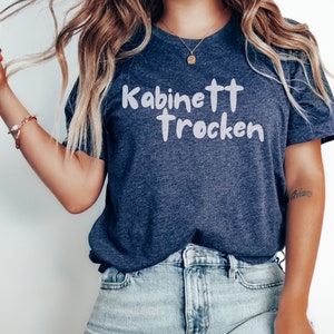 May include: A navy blue t-shirt with white text that reads "Kabinett trocken". The shirt is being worn by a person with long blonde hair and is paired with light blue jeans.