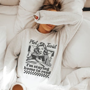 May include: A gray sweatshirt with a black and white illustration of a woman reading a book in bed. The text "Plot Twist I'm staying home to read" is printed on the sweatshirt.