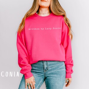 May include: A bright pink sweatshirt with the text "written by Lucy Score" in white.