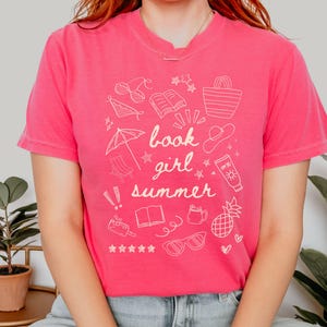 May include: A pink t-shirt with a white graphic design featuring the text "book girl summer" and illustrations of summer-themed items like sunglasses, a book, a beach umbrella, a pineapple, and a beach bag.