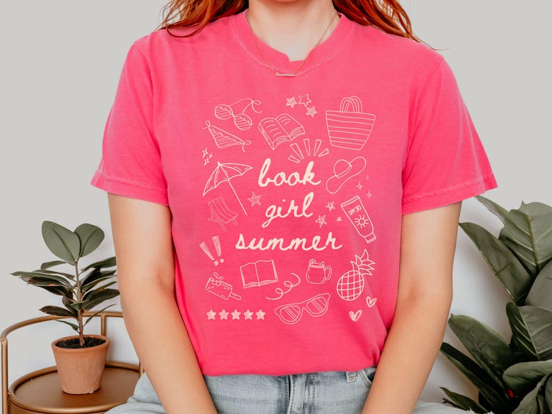 May include: A pink t-shirt with a white graphic design featuring the text "book girl summer" and illustrations of summer-themed items like sunglasses, a book, a beach umbrella, a pineapple, and a beach bag.