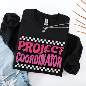 May include: Black sweatshirt with the words "PROJECT COORDINATOR" in pink, outlined in black, with a checkered pattern above and below the text. A silver necklace with a pendant is on the sweatshirt. The sweatshirt is on top of a pair of blue jeans.