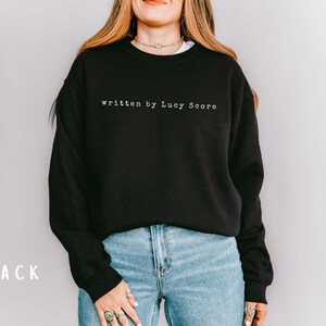 May include: Black sweatshirt with the text "written by Lucy Score" in white.