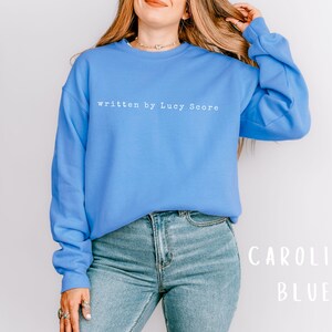 May include: A blue sweatshirt with the text "written by Lucy Score" in white. The sweatshirt is being worn by a person with long blonde hair. The background is white.