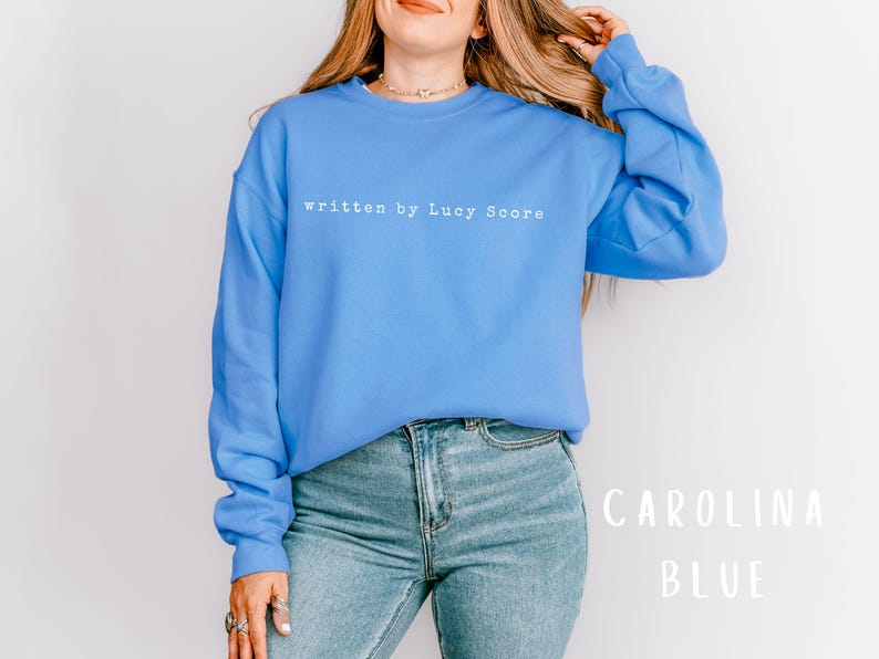 May include: A blue sweatshirt with the text "written by Lucy Score" in white. The sweatshirt is being worn by a person with long blonde hair. The background is white.