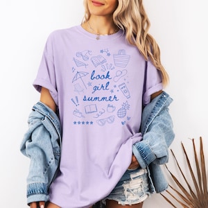 May include: A lavender t-shirt with a blue graphic design of summer-themed items and the text "book girl summer".