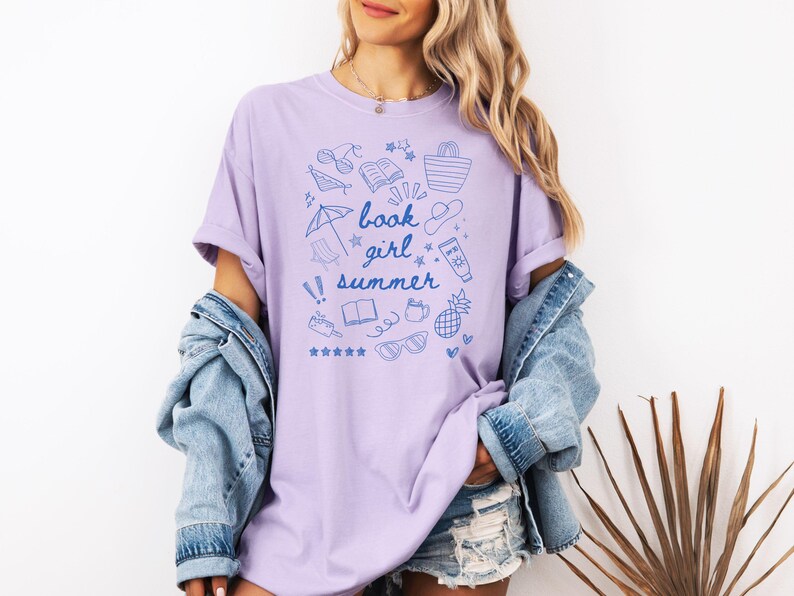 May include: A lavender t-shirt with a blue graphic design of summer-themed items and the text "book girl summer".