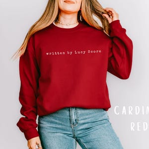 May include: A red sweatshirt with the text "written by Lucy Score" in white. The model is wearing blue jeans.