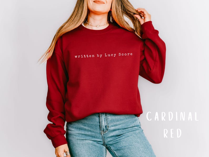 May include: A red sweatshirt with the text "written by Lucy Score" in white. The model is wearing blue jeans.