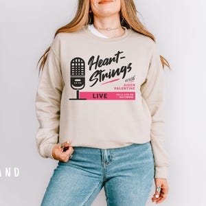 Puede incluir: Una sudadera beige con el texto "Heart-Strings with Aiden Valentine 103.1 Lite FM Baltimore LIVE" impreso en la parte delantera. El texto est&aacute; en negro y rosa.
