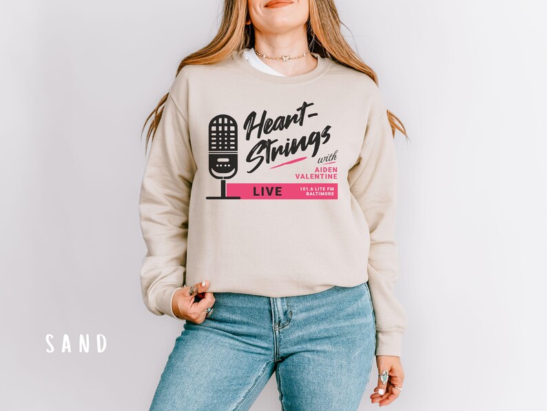 Puede incluir: Una sudadera beige con el texto "Heart-Strings with Aiden Valentine 103.1 Lite FM Baltimore LIVE" impreso en la parte delantera. El texto est&aacute; en negro y rosa.