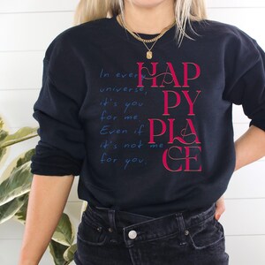 Może przedstawiać: Czarna bluza z nadrukiem "In every universe, it's you for me. Even if it's not me for you. Happy Place" w czerwonym i niebieskim kolorze.
