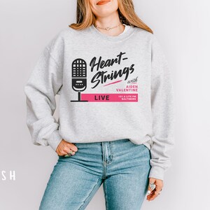 Puede incluir: Una sudadera gris con el texto "Heart-Strings with Aiden Valentine 101.3 Lite FM Baltimore LIVE" impreso en la parte delantera. El texto est&aacute; en negro y rosa.