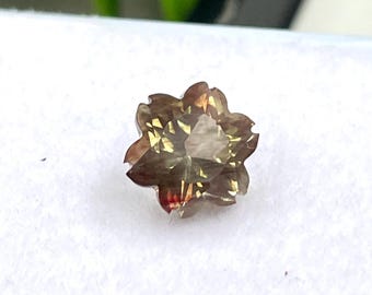 May include: A faceted gemstone with a flower-like shape. The gem features a mix of brown, gold, and red hues. The gemstone is set against a white background.