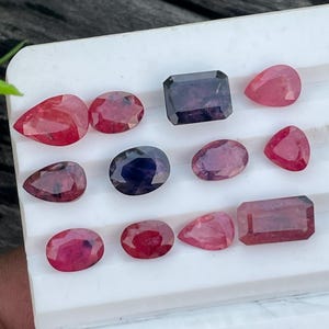 Op de afbeelding: Assortiment van gepolijste edelstenen in verschillende vormen en tinten rood, roze en paars. De edelstenen worden getoond op een wit oppervlak, met een donkere houten achtergrond. Inclusief ovale, peervormige en rechthoekige slijpsels.