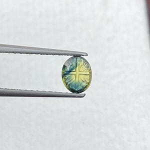 May include: An oval-shaped gemstone featuring a distinct cross pattern carved into its surface. The stone displays a gradient of colors, transitioning from deep blue at the edges to a bright yellow center, held by a pair of metal tweezers.