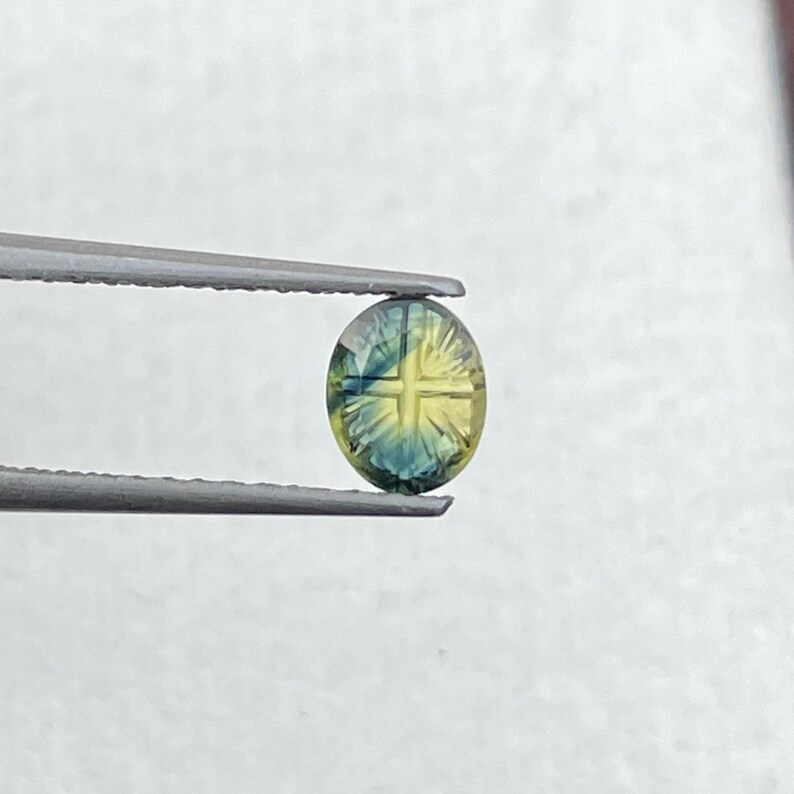 May include: An oval-shaped gemstone featuring a distinct cross pattern carved into its surface. The stone displays a gradient of colors, transitioning from deep blue at the edges to a bright yellow center, held by a pair of metal tweezers.